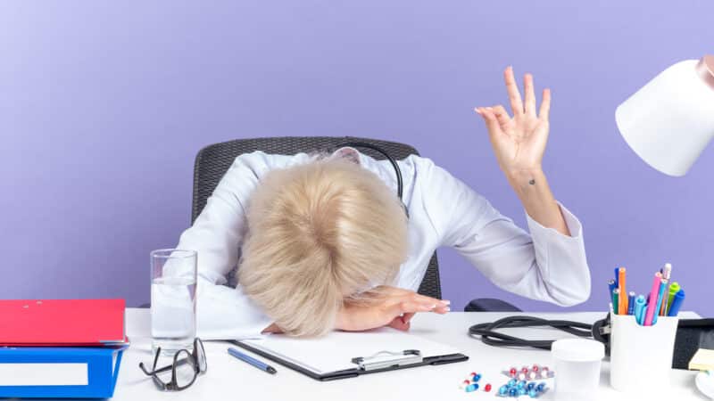 tired adult slavic female doctor in medical robe with stethoscope sitting at desk with office tools putting head on desk and gesturing ok sign isolated on purple background with copy space